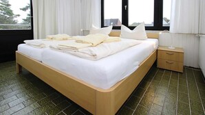 Uma cama bem arrumada com lençóis e almofadas brancas, uma mesa de cabeceira com abajur e uma janela com cortinas.
