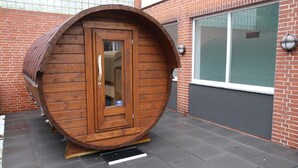 Uma estrutura em forma de barril de madeira, com uma porta, situada sobre um piso pavimentado, com um edifício de tijolos ao fundo.