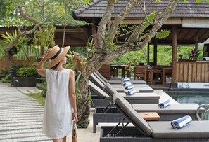 Una mujer con un vestido blanco de pie junto a una zona de lounge junto a la piscina, con tumbonas y un área de estar cubierta.
