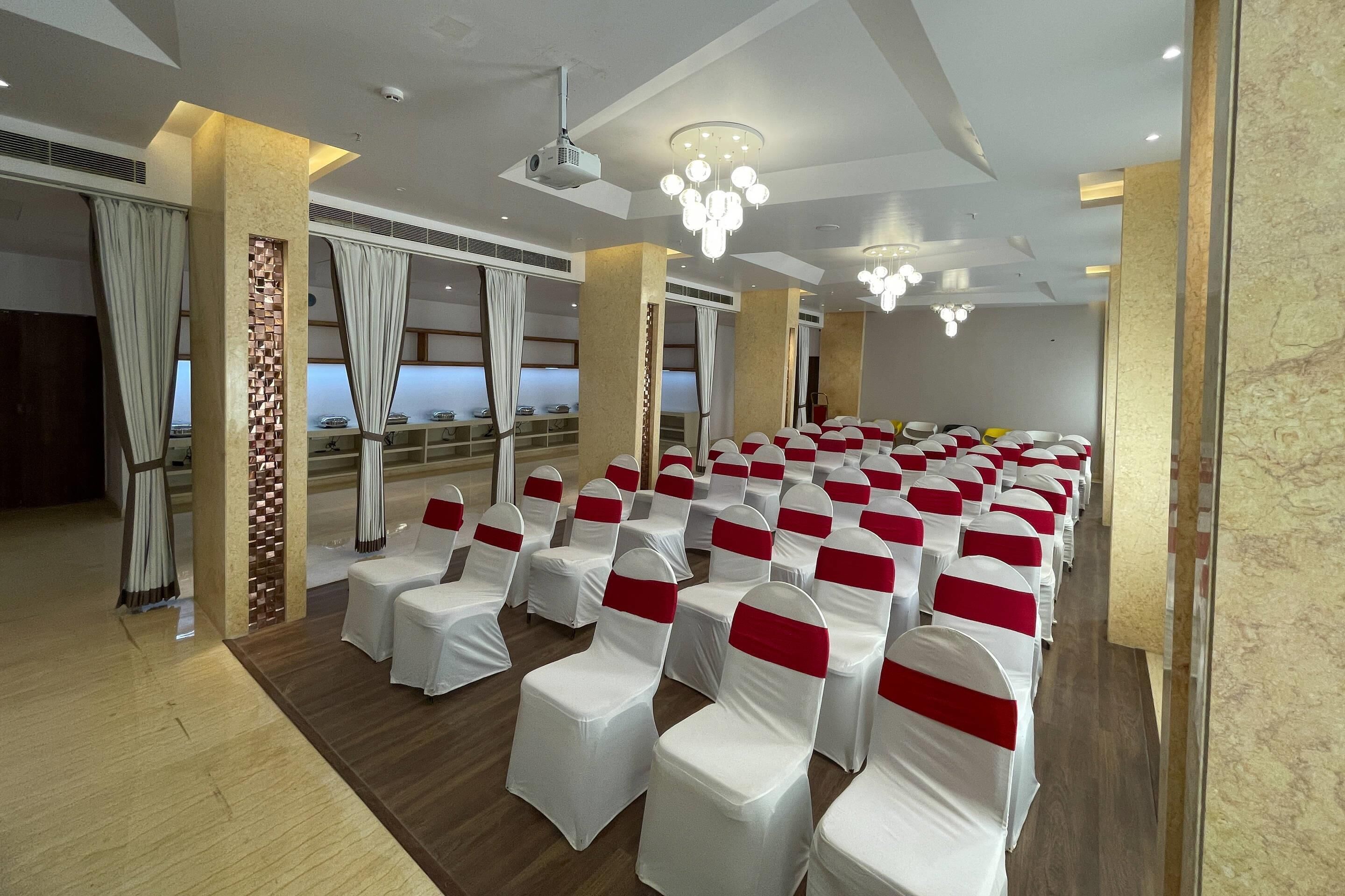 A conference room with rows of chairs covered in white and red sashes, a marble wall, and a ceiling with recessed lighting and a projector.