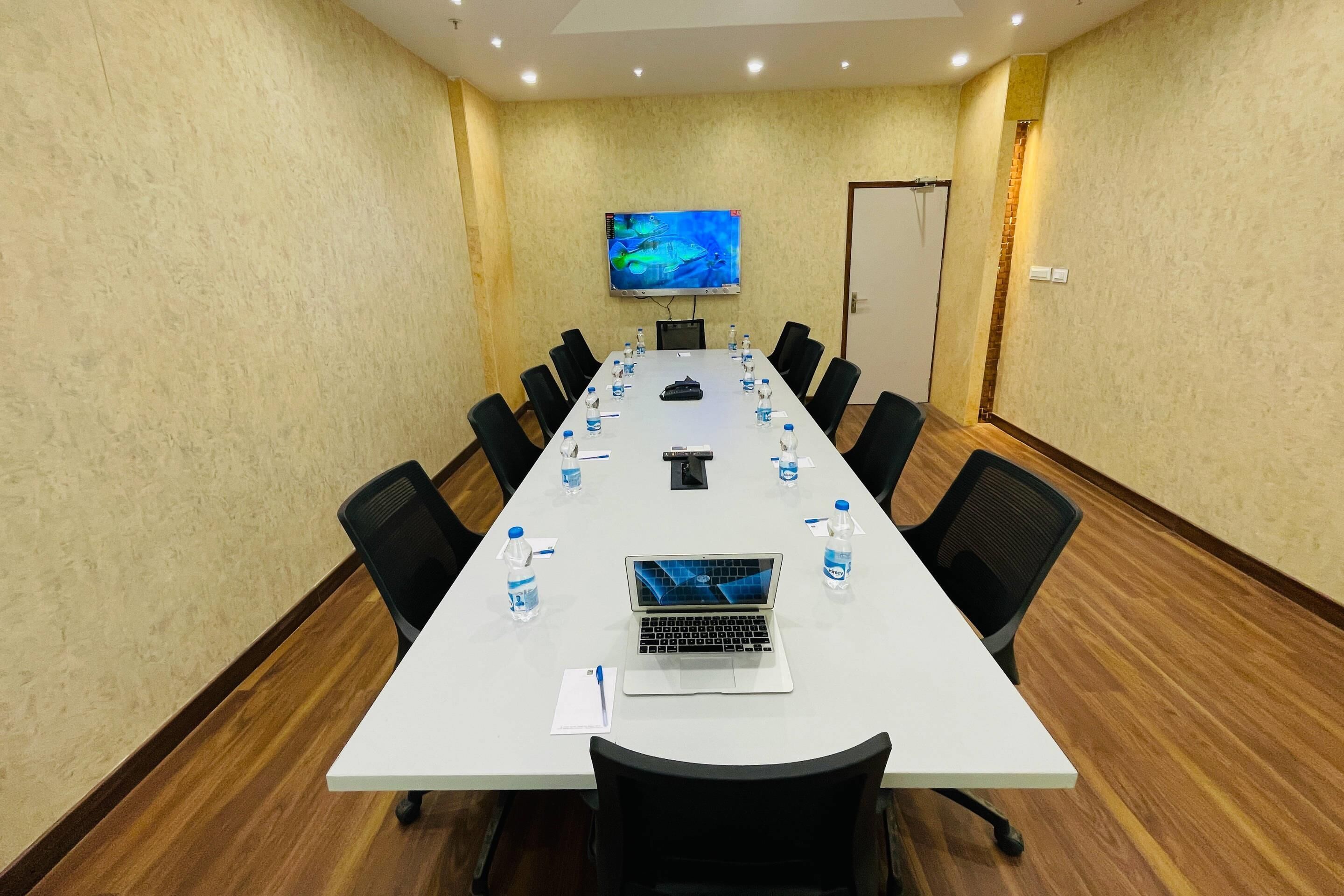 A conference room with a long table, black chairs, a laptop, water bottles, and a TV on the wall.