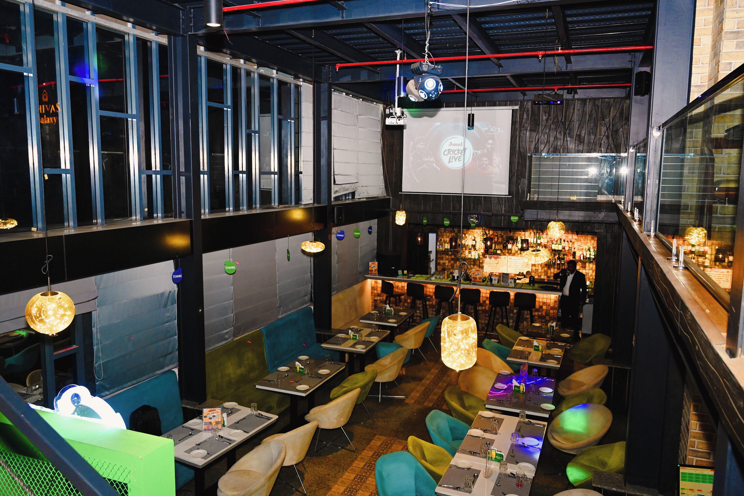 A restaurant with multiple dining areas, a bar, and a projection screen displaying 'Annual Cricket Live'.