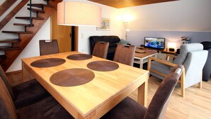 A modern living room with a wooden dining table, brown chairs, a sofa, a TV, and a staircase. - Greetjehaus mit Garten, Nähe Strand, Dornumersiel (Dornumersiel)