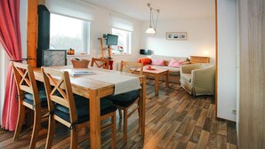 A cozy living room with a wooden dining table, chairs, and a sofa. There is a fireplace, a television, and a window with curtains. - Ferienwohnungen am Plauer See, Zislow (Zislow)