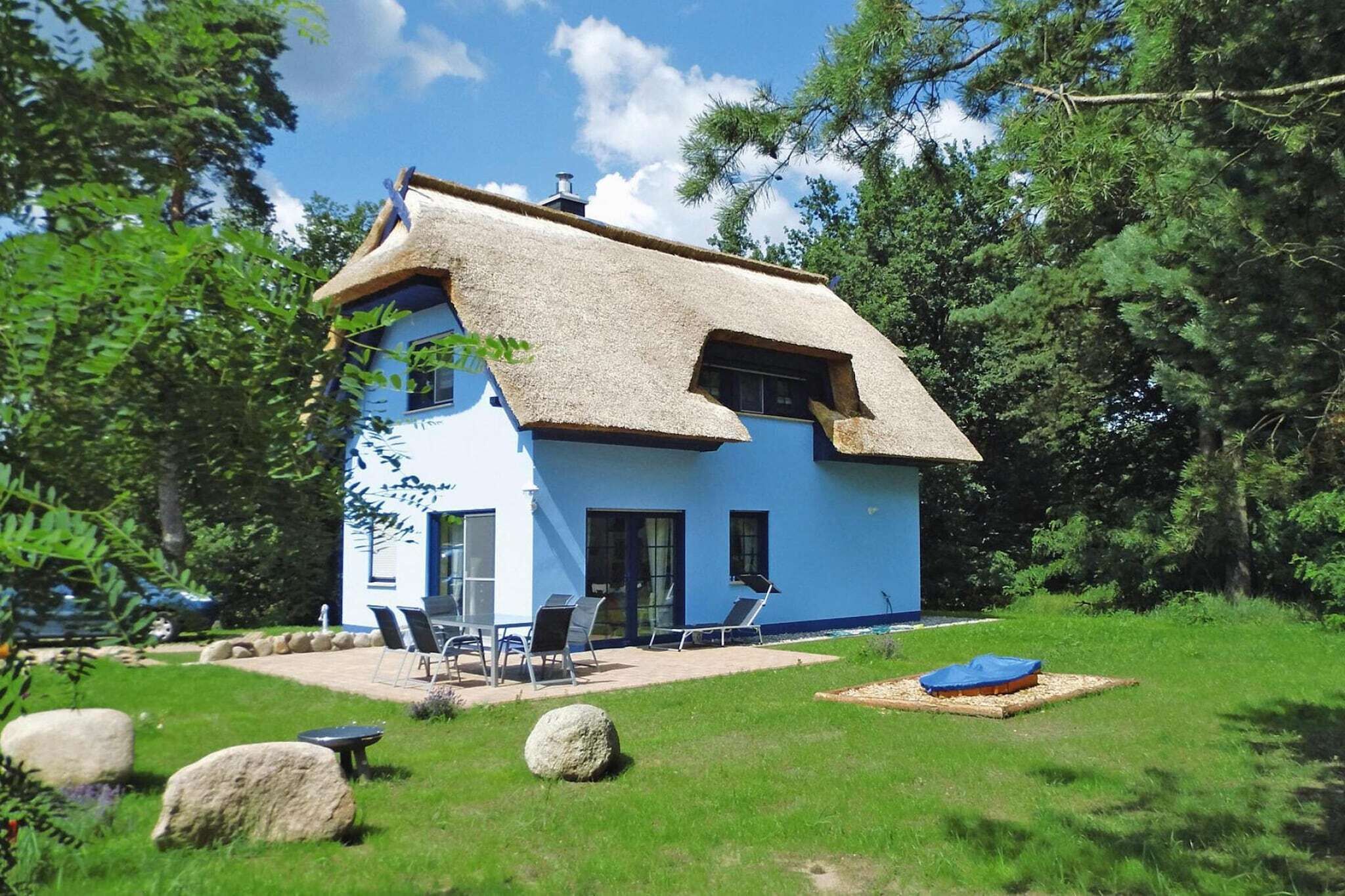 Ein zweistöckiges Haus mit Strohdach, blauer Fassade und einer Terrasse mit Gartenmöbeln.