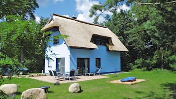 Ein zweistöckiges Haus mit Strohdach, blauer Fassade und einer Terrasse mit Gartenmöbeln.