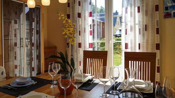 A dining area with a table set for four, a vase with yellow flowers, and a view of the outdoors through large windows. - Ferienhaus Nachtigall, Glowe (Glowe)
