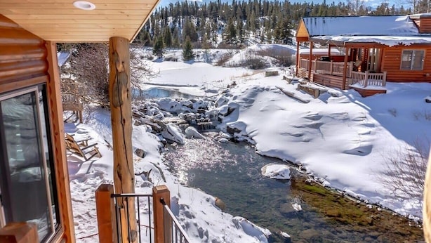 Une cabane en bois dotée d’une terrasse offrant une vue sur un paysage enneigé et un ruisseau aux eaux claires.