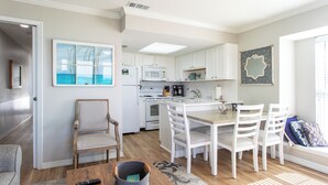 Une cuisine moderne avec des meubles blancs, un four à micro-ondes et une cuisinière. On y trouve un coin repas avec une table en bois et des chaises blanches. Une fenêtre laisse entrer la lumière naturelle.
