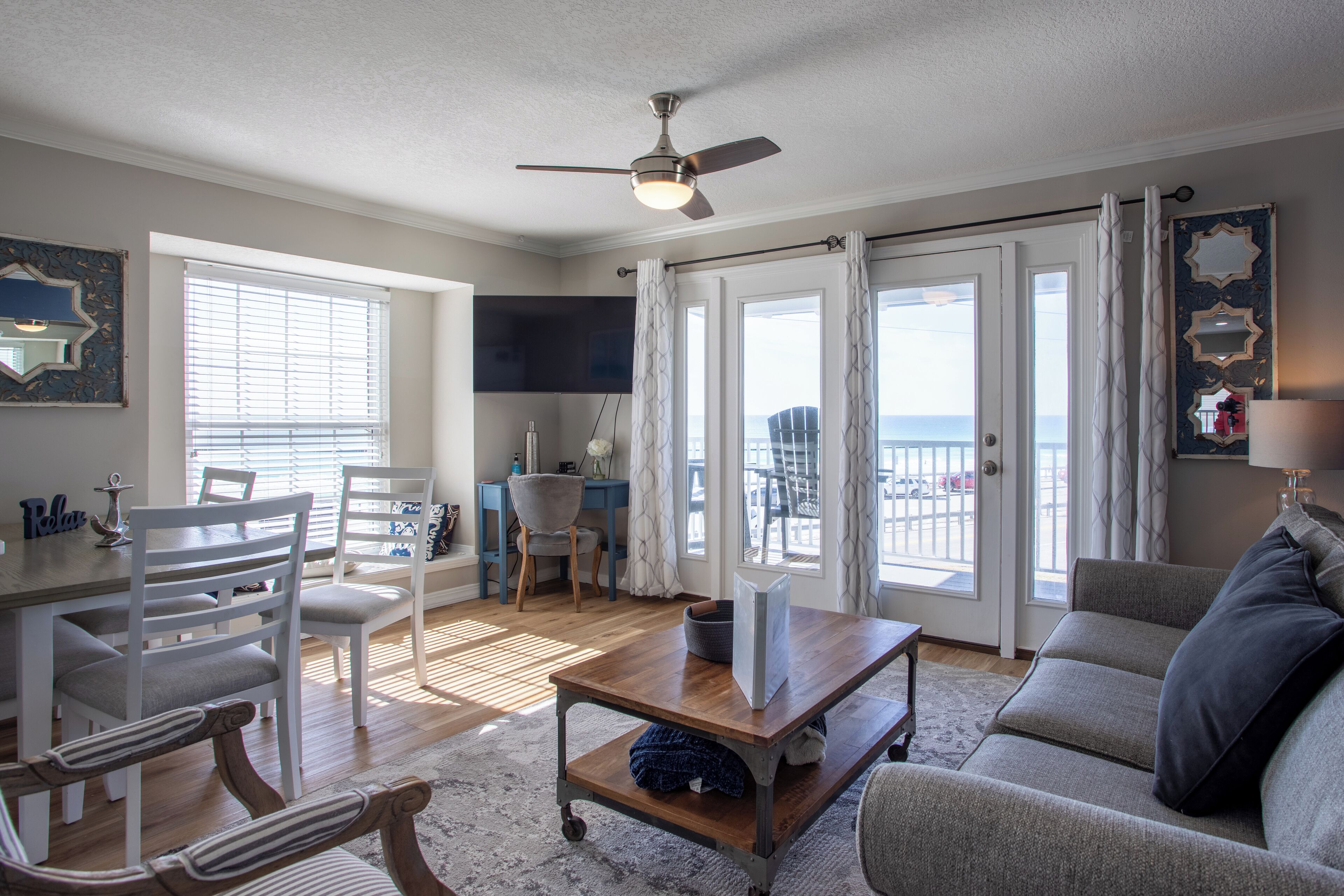 A spacious living room with a ceiling fan, a dining area with white chairs, a sofa, and a view of the ocean through large windows.