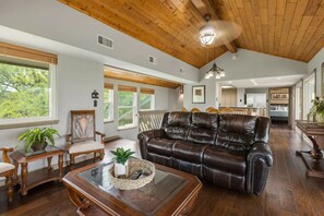 A spacious living room with a leather sofa, wooden furniture, and large windows offering a view of the outdoors.