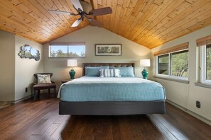 A bedroom with a large bed, wooden ceiling, two bedside lamps, a chair, and a window with a view.