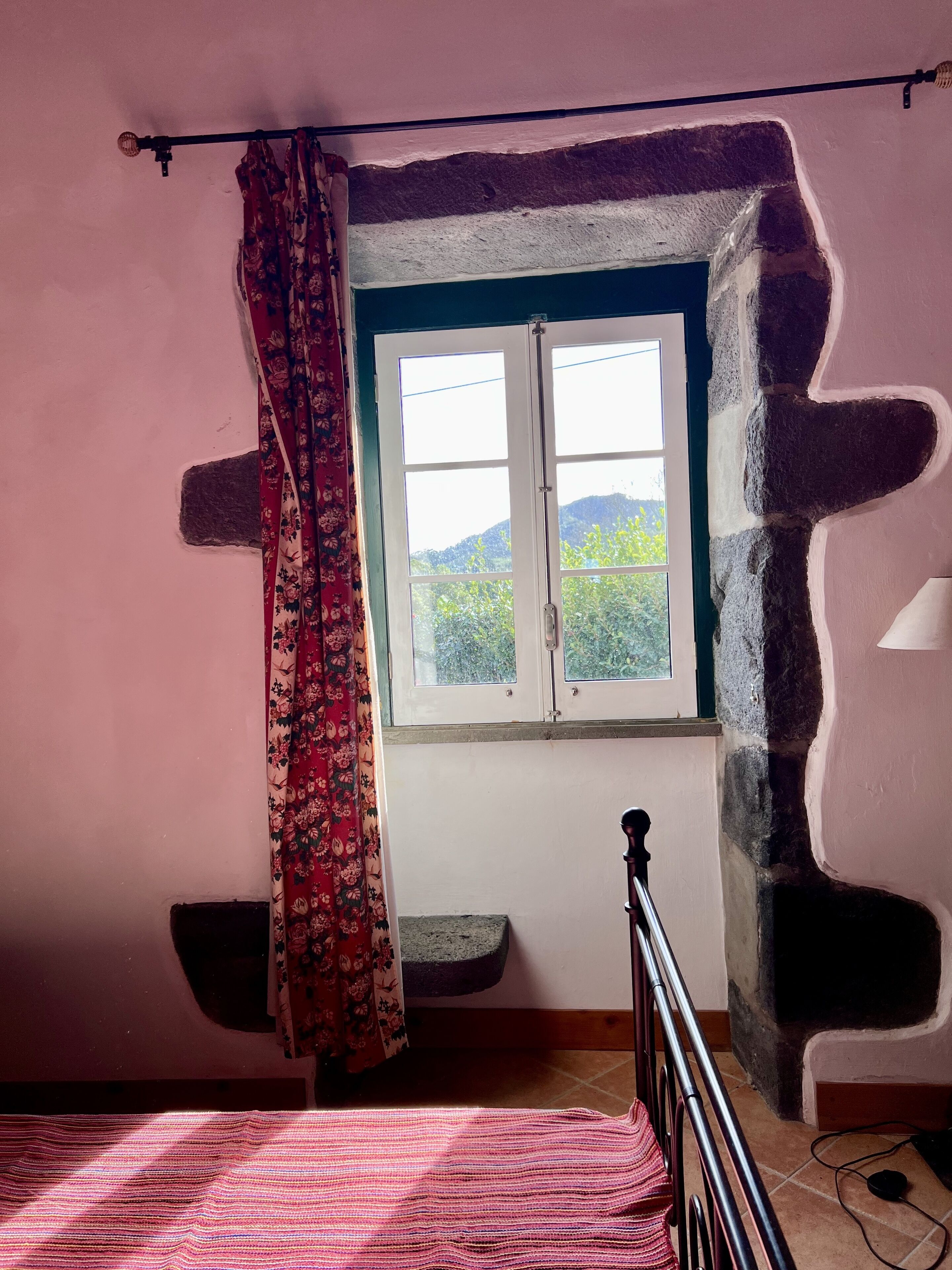 A bedroom with a bed, a window with a view of greenery, a curtain, and a stone wall.