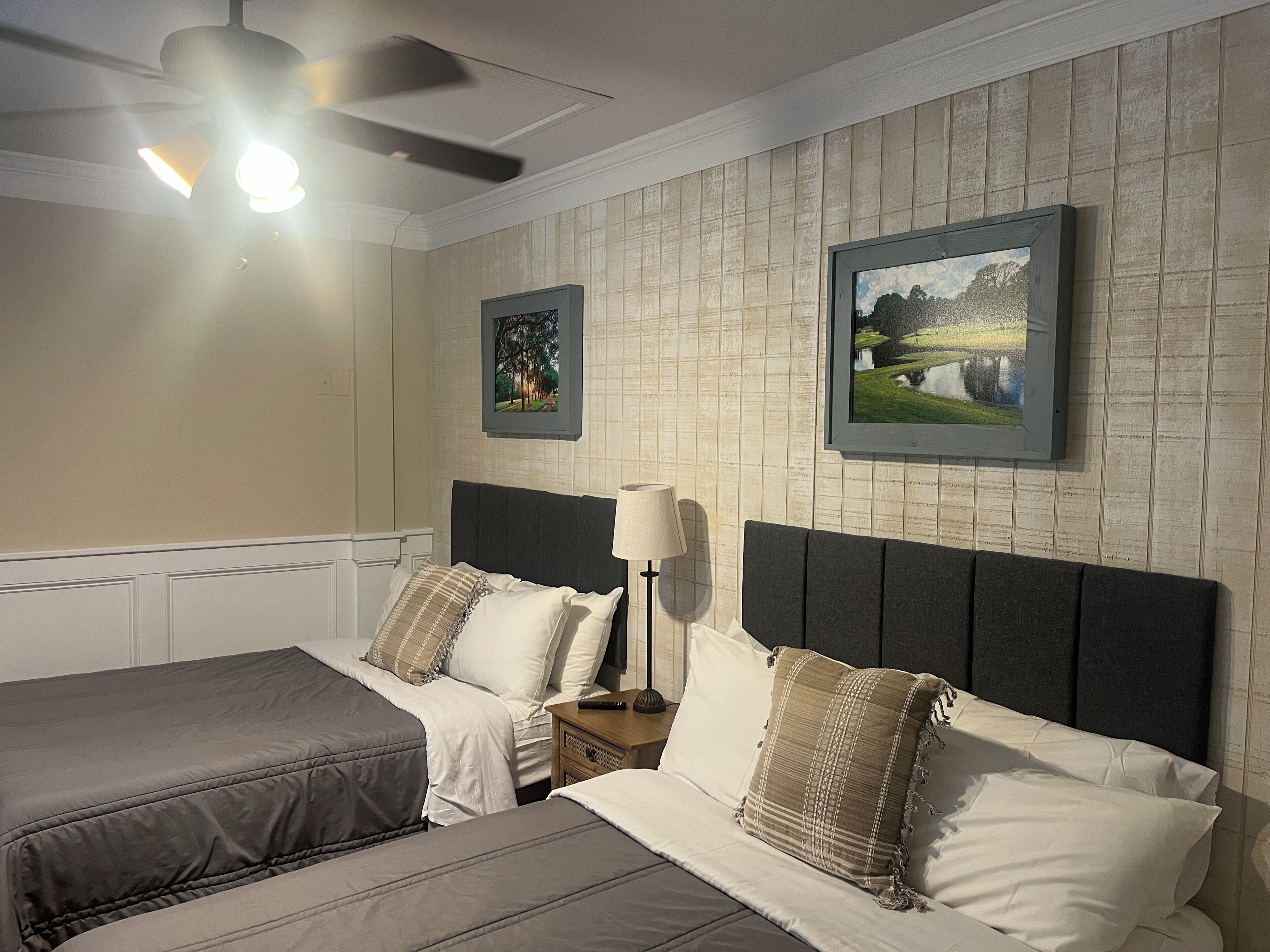 Een hotelkamer met twee bedden, een plafondventilator, een nachtkastje met een lamp en twee ingekaderde foto's aan de muur.