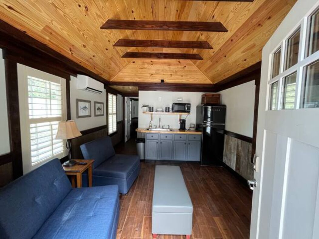 Una stanza accogliente con soffitto in legno, un divano, una panca, un angolo cottura con pensili ed elettrodomestici, e una porta con vetri.