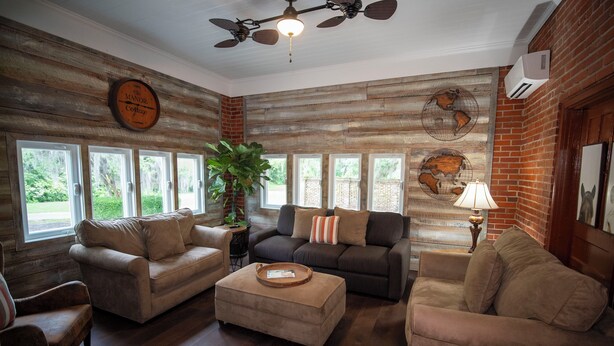 A cozy living room with wooden walls, a ceiling fan, and a large window overlooking greenery. - Wedgefield Country Club  (Georgetown)