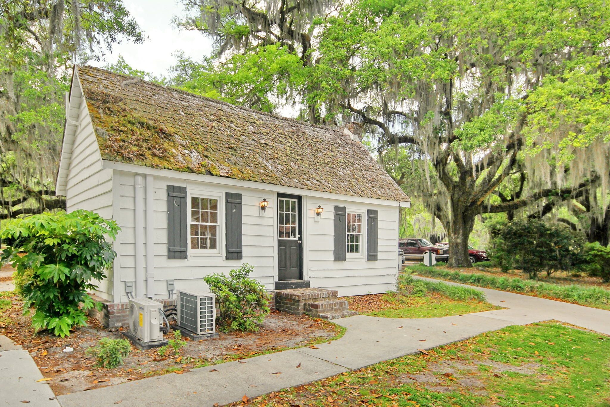 Une petite maison blanche avec un toit en pente, entourée d’arbres et bordée d’un trottoir.