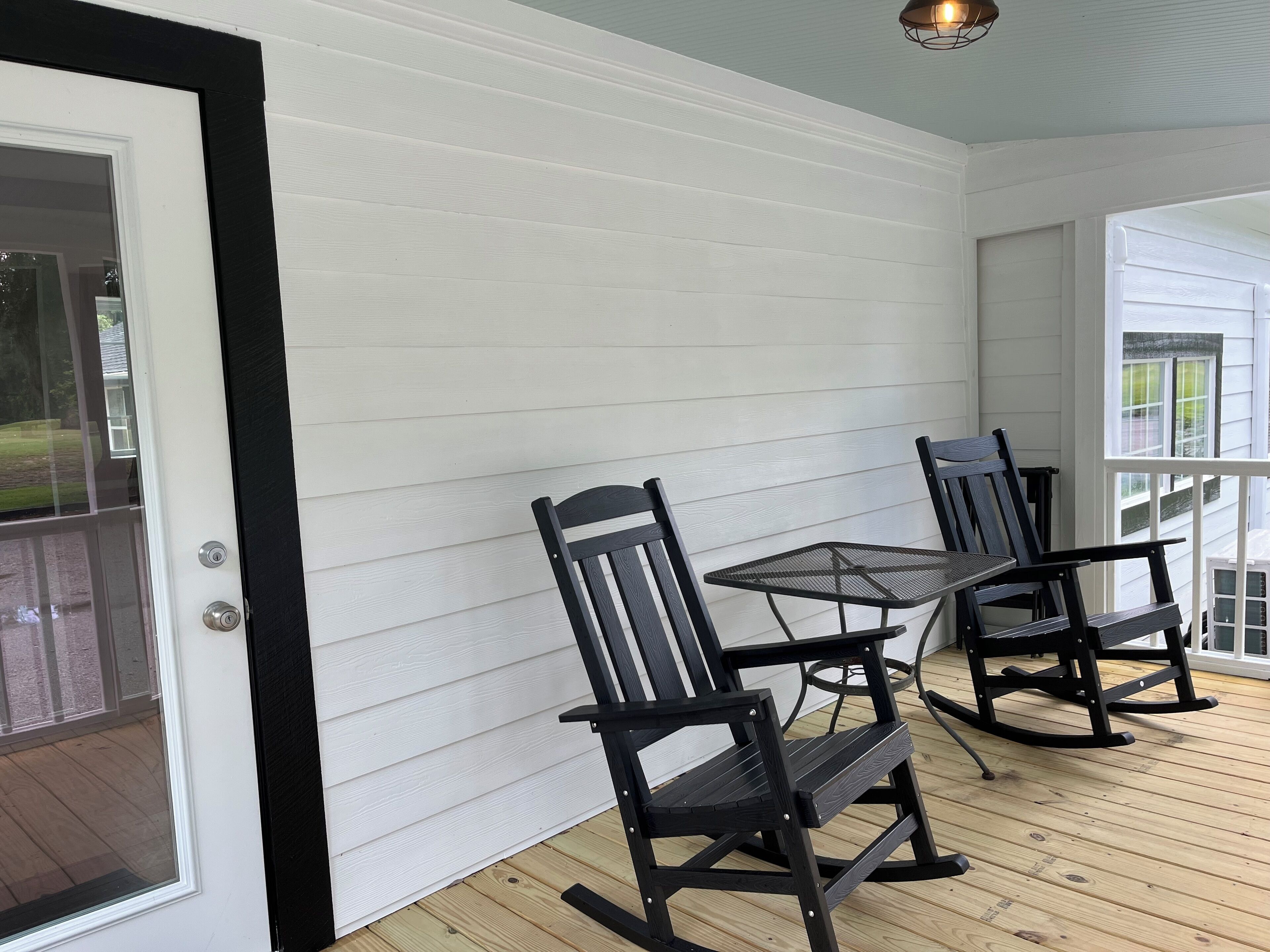 A covered patio with a wooden deck, white siding walls, and black furniture including chairs and a table.