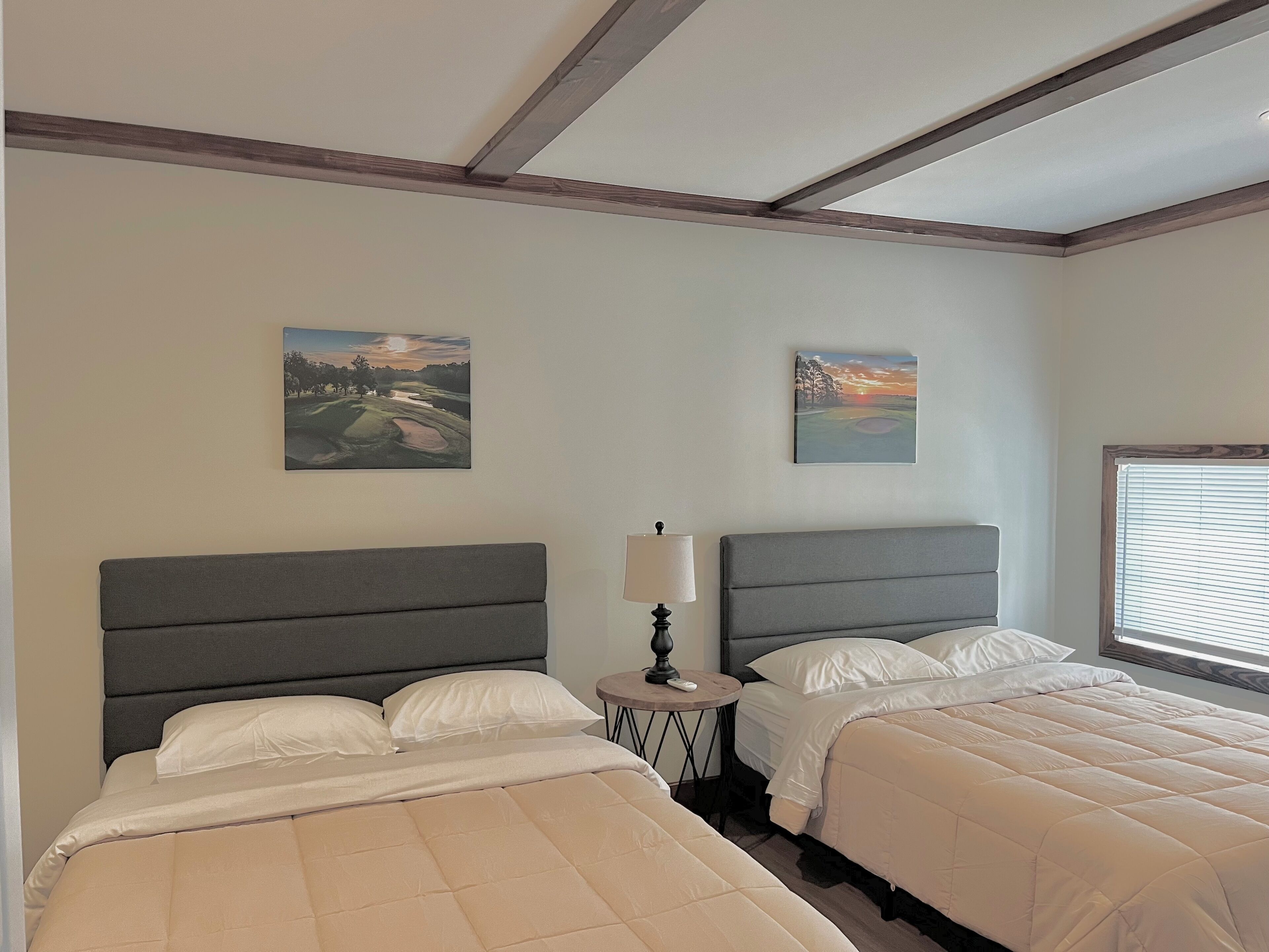 A hotel room with two beds, a round table with a lamp, and two paintings on the walls.