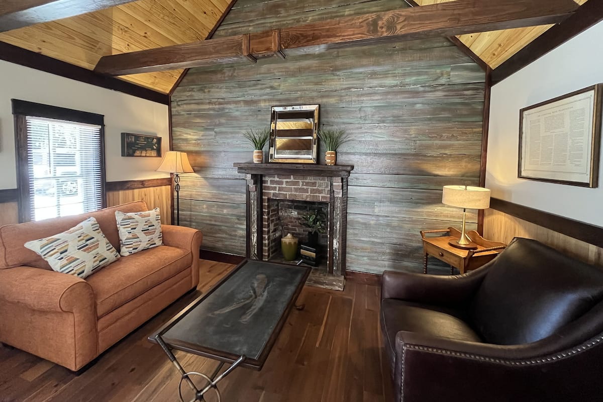 A cozy living room with a fireplace, wooden beams, a sofa, a coffee table, and a leather armchair.
