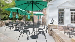 An outdoor patio with tables and chairs, green umbrellas, and a white building with a brick chimney.