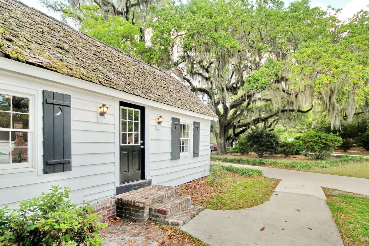 A white building with a black door and windows, surrounded by lush greenery and a tree with hanging moss.