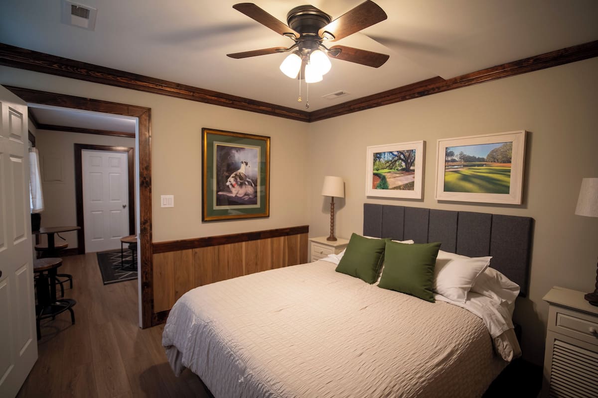 A bedroom with a large bed, a ceiling fan, a nightstand with a lamp, and framed pictures on the wall.