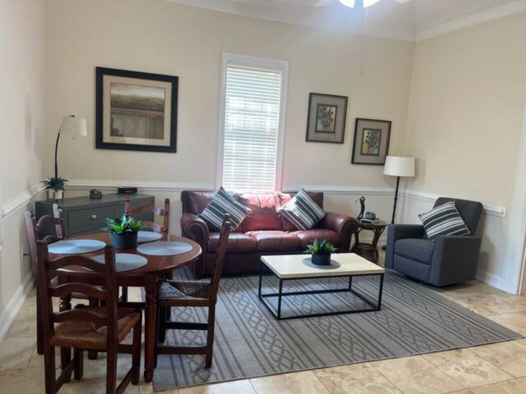 A living room with a brown leather sofa, a gray armchair, a coffee table, and a dining table with chairs. There are framed pictures on the wall and a lamp on a side table.