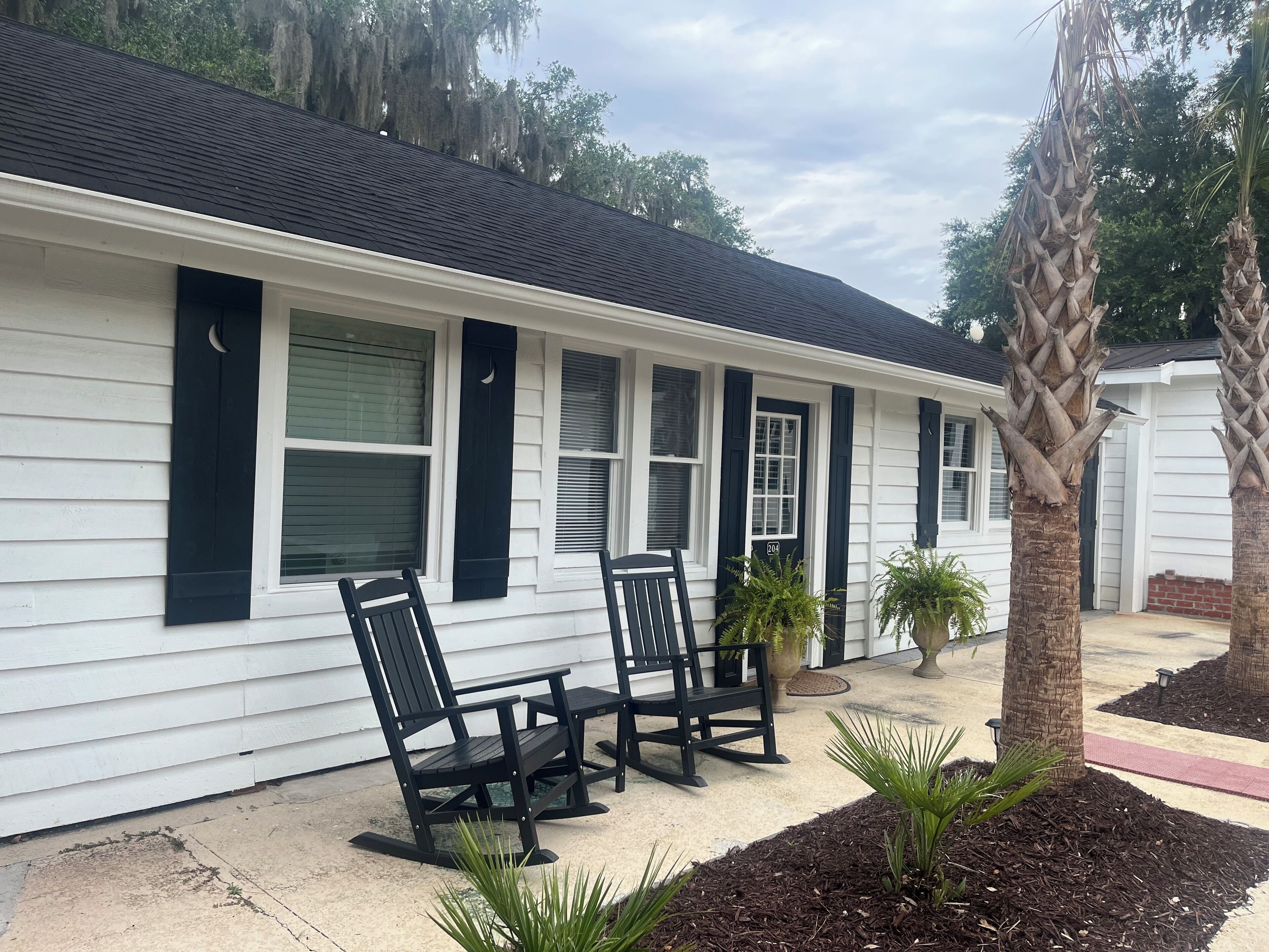 A white building with black shutters, two rocking chairs, and a palm tree.