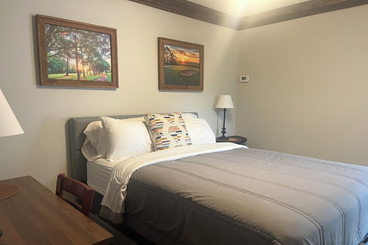 A neatly made bed with a grey duvet, a pillow with a colorful pattern, and two framed landscape paintings on the wall.