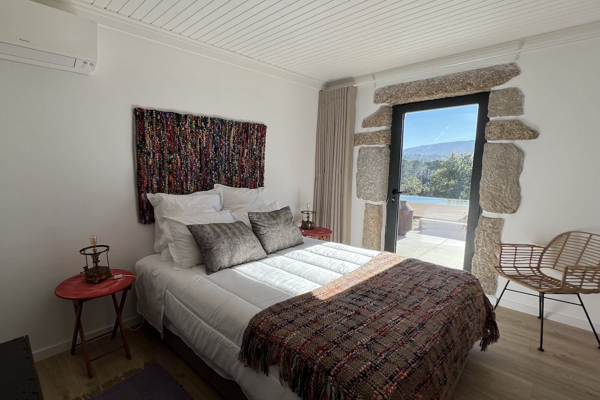 Un dormitorio con una cama, una mesita de noche roja, una manta decorativa estampada, un adorno en la pared y vistas a través de un ventanal.