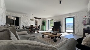 Amplia sala de estar con sofá, una mesa de centro y televisión. Cuenta con un comedor con sillas y mesa. La habitación tiene una pared de piedra y un ventanal grande.