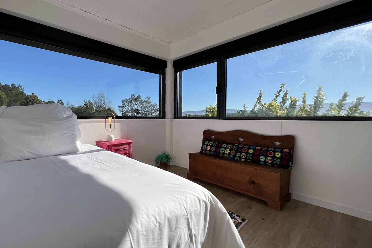 Un dormitorio con cama, mesita de noche, banco de madera y ventanas amplias que ofrecen vistas a árboles y montañas.