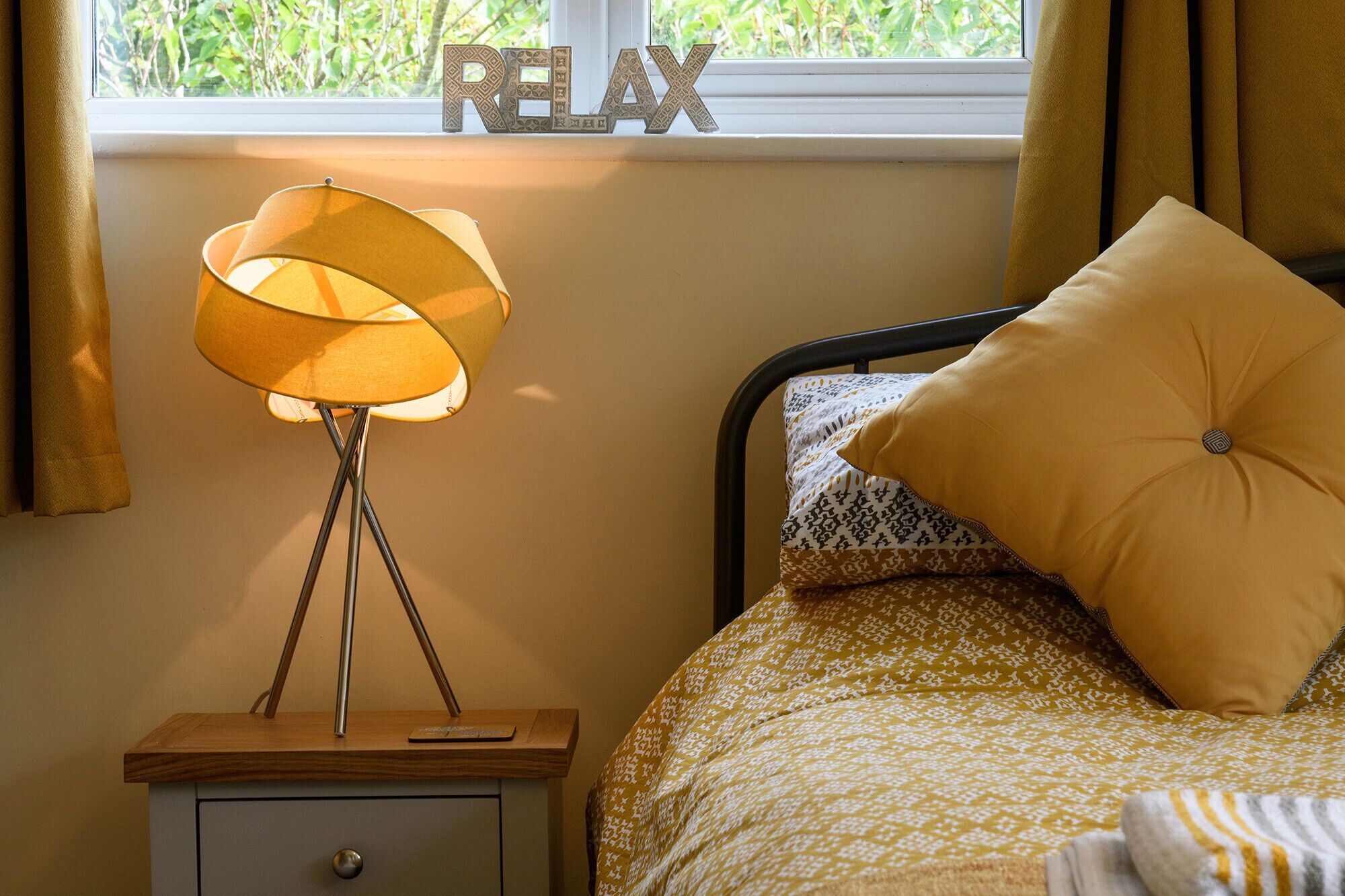 A bedroom with a bed, a bedside table with a lamp, a window with curtains, and a 'RELAX' sign on the wall.