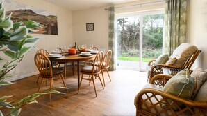 A dining area with a round wooden table, four chairs, and a dining set. There is a large window with curtains, a view of greenery outside, and a painting on the wall. - Cosy family cottage close to Beddgelert and Porthmadog (Porthmadog)