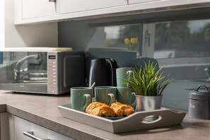 Eine Küchenarbeitsplatte mit einer Mikrowelle, einem Wasserkocher, einer Platte mit Croissants und einer Topfpflanze.