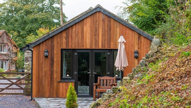 A wooden cabin with a glass door, a small patio with a chair and table, and a stone wall.