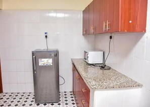 A compact kitchen with white tiled walls, a granite countertop, a refrigerator, and a microwave.