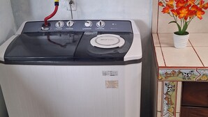 A compact, white washing machine with a red hose and a control panel, situated in a corner of a room with tiled walls and a wooden cabinet. - Bosque del Río Lodging (Monteverde)