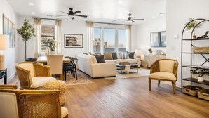 A spacious living room with a sofa, chairs, a coffee table, and a dining area. There are ceiling fans, large windows, and a view of the city. - Shoreline: Desert Color Lagoon + Beaches (St. George)