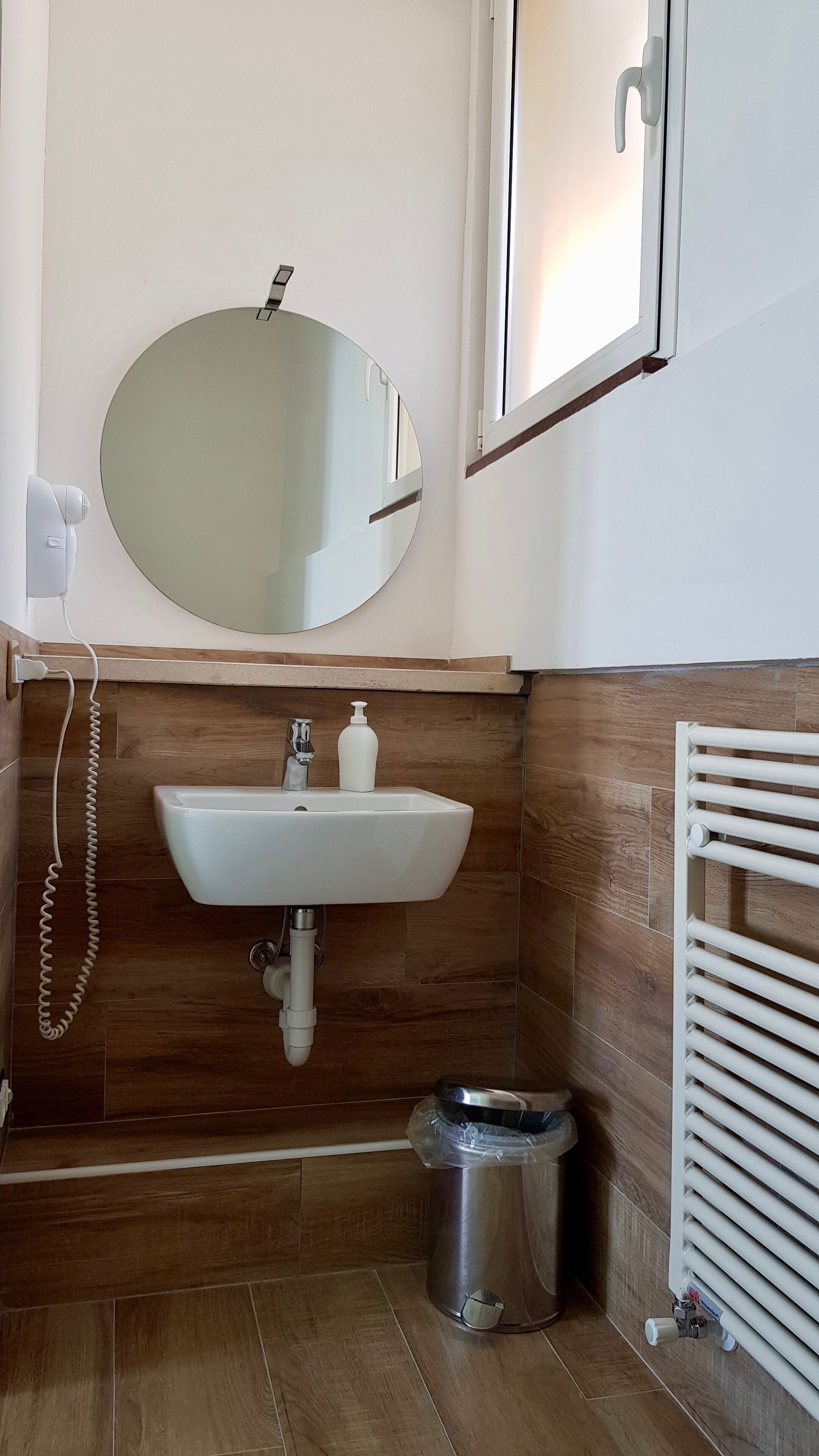 A bathroom with a white sink, wooden wall paneling, a mirror, a radiator, and a trash bin.