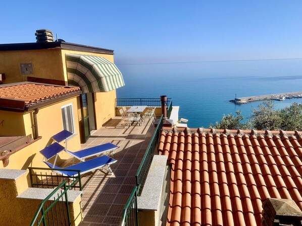 A balcony with a table and chairs, overlooking a coastal area with a clear view of the sea and a small pier. - Beautiful panoramic terrace with a view of the ocean and a swimming pool (Finale Ligure)