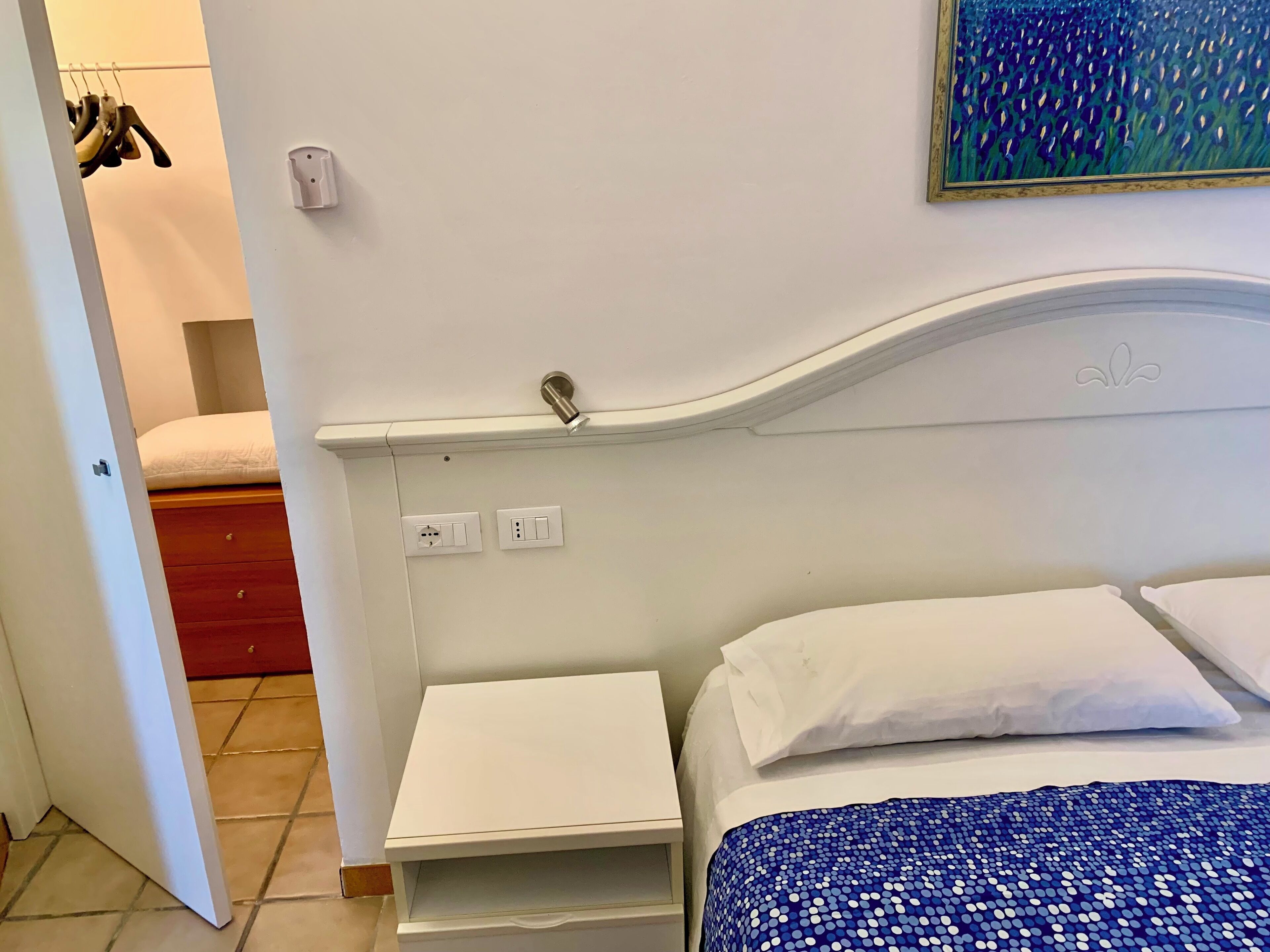 Ein Schlafzimmer mit Bett, Nachttisch, kleinem Tisch und einer Wandleuchte. Eine Tür führt in einen anderen Raum, ein Bild ist an der Wand.