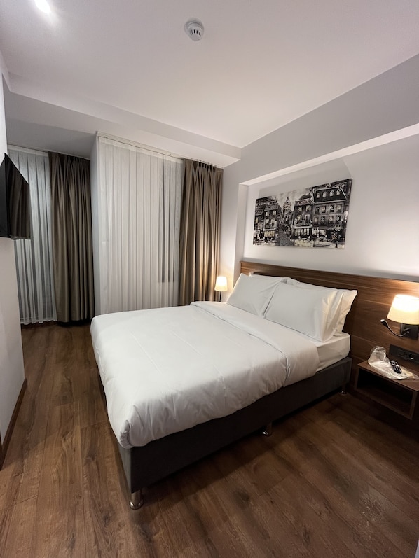 A hotel room with a large bed, wooden flooring, a mounted TV, a painting on the wall, and a nightstand with a lamp and a remote control. - PALMERA HAGIA SOPHIA (Istanbul)
