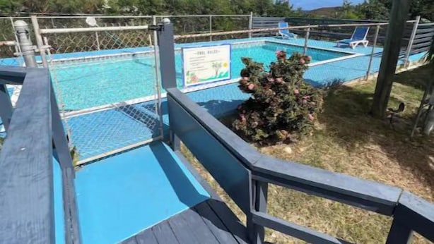 Une terrasse en bois mène à un espace piscine agrémenté d’un panneau indicateur, le tout étant entouré de verdure et surplombé par un ciel dégagé.