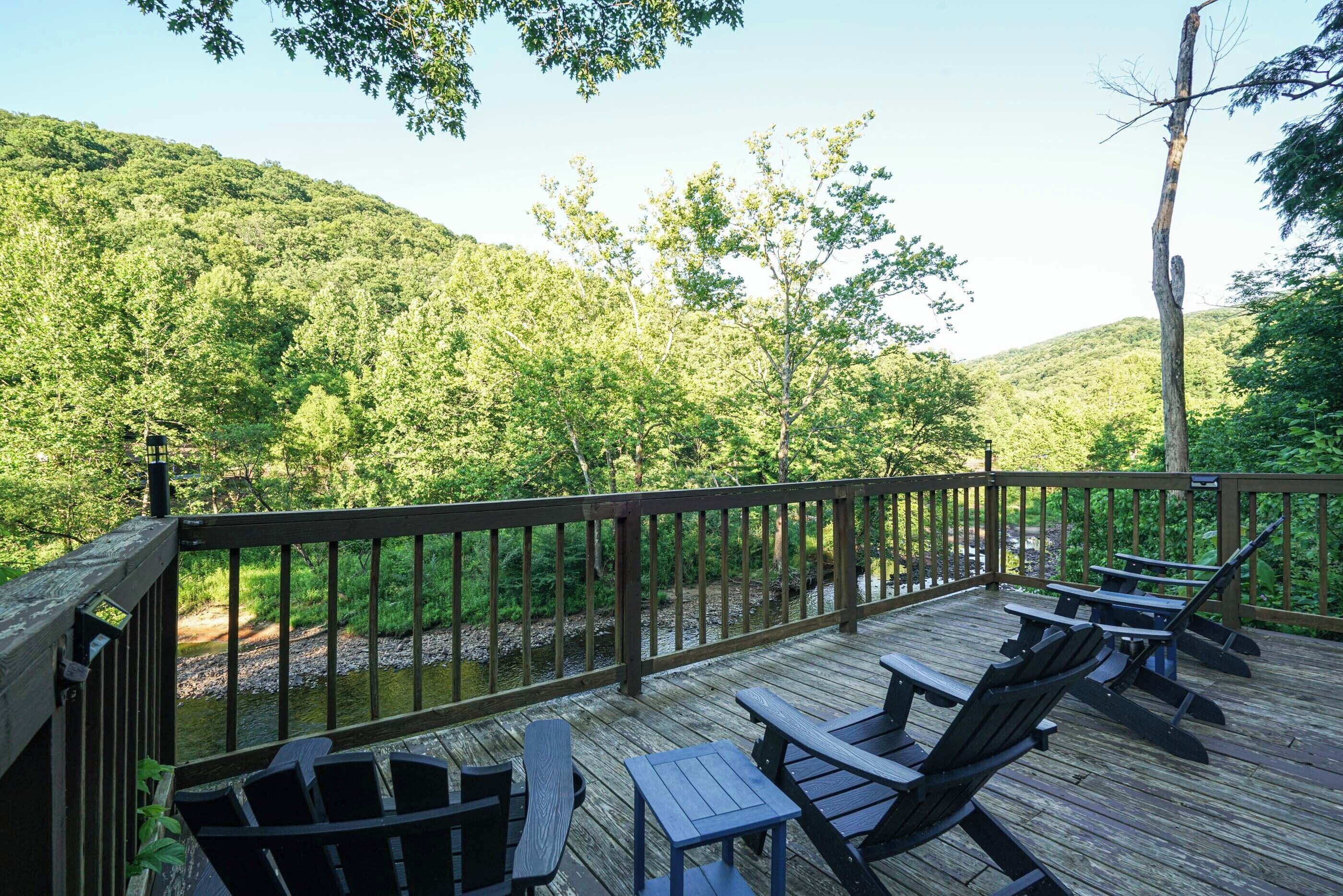 A wooden deck with picnic tables and chairs, overlooking a river and forested hills.