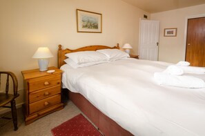 A neatly made bed with white linens, a wooden nightstand with a lamp, a red rug, and a framed picture on the wall.