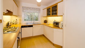 A compact kitchen with a stove, sink, and microwave. It features white cabinets, a wooden countertop, and a window with a view of trees. - 18 Thrang Brow - Two Bedroom House, Sleeps 4 (Ambleside)