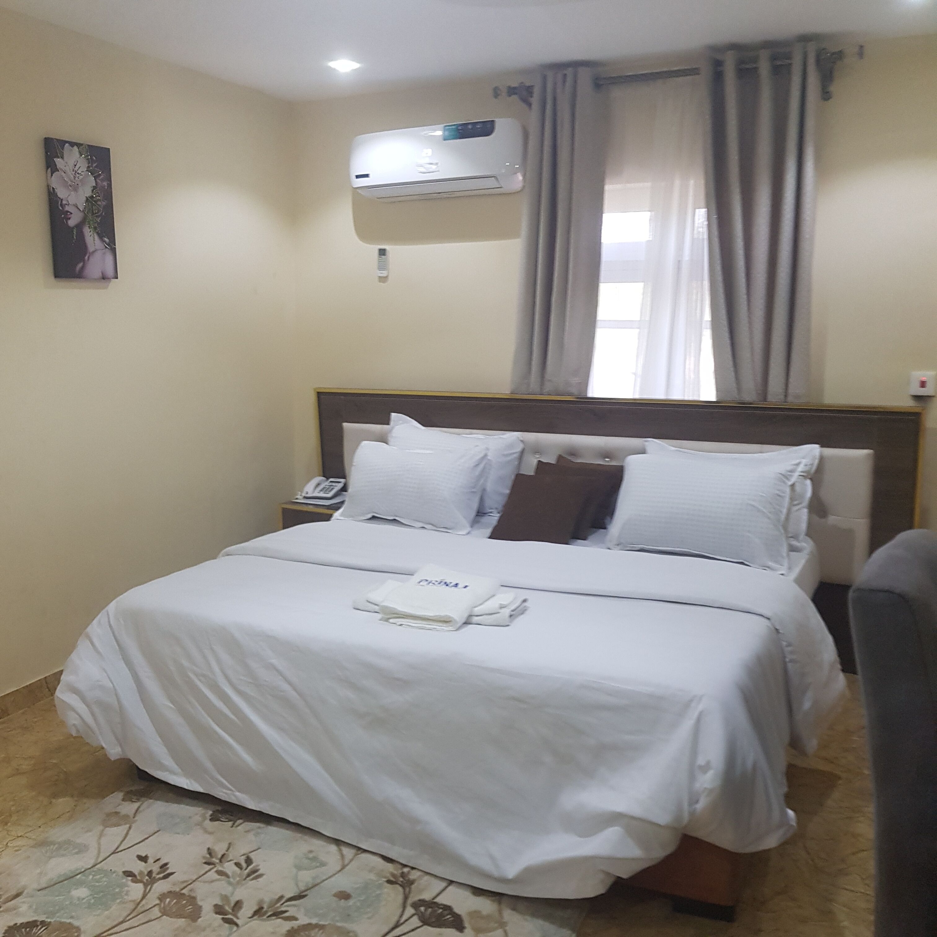 A hotel room with a bed, a bedside phone, and an air conditioning unit.
