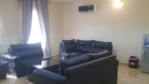 A living room with a black leather sofa, a black armchair, a small table, a TV mounted on the wall, and a refrigerator.